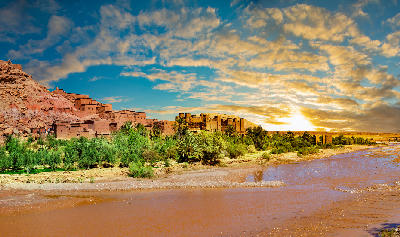 Sunset view of Aït Benhaddou in Morocco with the ancient kasbah village, palm oasis, and riverbed in the foreground.