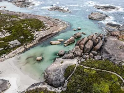 High-angle aerial view of a cluster of giant, rounded granite boulders sitting in a shallow, crystal-clear turquoise cove next to a white sand beach and coastal vegetation.