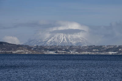 Snow-covered Mount Usu rising beyond Lake Toya with low clouds wrapping the summit, viewed across the lake in Hokkaido, Japan.