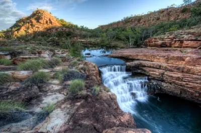 Water cascades over layered red rock into a deep pool surrounded by rugged cliffs and native vegetation at Bell Gorge.
