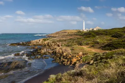 Cape Leeuwin Lighthouse rises above a rocky shoreline and coastal vegetation beside the ocean.