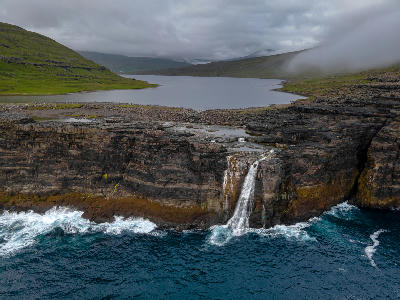 Aerial view of a visitor overlooking Bøsdalafossur waterfall as it drops from Lake Sørvágsvatn into the Atlantic, with the Trælanípa cliffs rising above the western coast of Vágar Island, Faroe Islands.