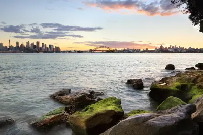 Sunset view from the rocky shoreline at Bradleys Head across Sydney Harbour towards the CBD, Opera House and Harbour Bridge, with moss-covered rocks in the foreground.