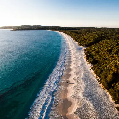Two visitors walk along the sweeping white sands of Hyams Beach beside clear blue water in warm early-morning light.
