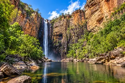 Tall waterfall descending into a reflective plunge pool beneath towering sandstone cliffs and green vegetation.