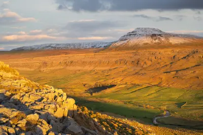 Snow-capped Ingleborough glowing in golden evening sunlight above limestone rocks and a winding river valley in the Yorkshire Dales, England.