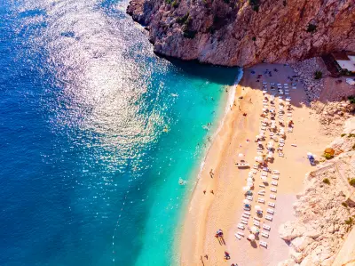 Dramatic aerial view of Kaputaş Beach, where brilliant turquoise Mediterranean water meets a small golden cove enclosed by steep rocky cliffs, with swimmers, sun loungers and umbrellas along the shore.