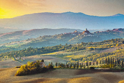 This breathtaking panoramic landscape captures the rolling hills of Tuscany, Italy, bathed in the soft, warm light of either sunrise or sunset. In the distance, perched atop a prominent hill, stands the imposing Rocca di Tentennano, a medieval fortress. The charming village of Castiglione d’Orcia clusters along a ridge next to it. The foreground and middle ground feature the iconic Tuscan scenery: gently undulating fields in hues of golden brown and green, interspersed with olive groves, and slender cypress trees. Distant hazy mountains form a picturesque backdrop under a sky with subtle gradients of color.