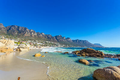 Wide view of Clifton Beach in Cape Town showing shallow transparent waves, rounded granite rocks on white sand and the Twelve Apostles mountains rising behind the Atlantic Seaboard.