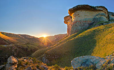 Sunset illuminating the towering Brandwag Buttress sandstone cliff above a grassy valley in Golden Gate Highlands National Park, with sun rays spreading across the surrounding mountain landscape.