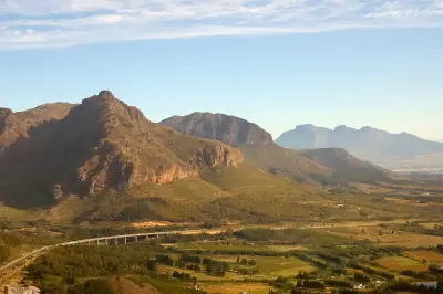 Mountain landscape seen from Du Toitskloof Pass near Paarl, South Africa, with winding highway, agricultural valleys, and layered Klein Drakenstein mountain ridges under soft afternoon light.