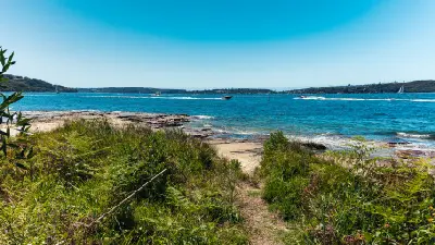 Rocky shoreline and native vegetation at Bradleys Head overlooking the blue waters of Sydney Harbour, with boats crossing the harbour in the distance.
