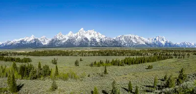 Panoramic view of the Grand Teton Range with snow-capped peaks above flat valley and scattered trees in Wyoming.