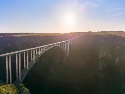 Aerial view of Bloukrans Bridge spanning a deep forested gorge at sunset on the Garden Route in South Africa, with the bridge silhouetted against the low sun above the canyon.