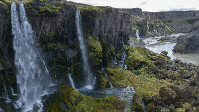 Aerial view of several waterfalls flowing down a tall stone cliff in the Iceland Highlands, with moss-covered rocks and a river winding through the valley.