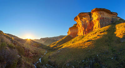 Sunrise illuminating the massive sandstone cliffs of Golden Gate Highlands National Park, with a small stream winding through a grassy mountain valley in the Free State, South Africa.