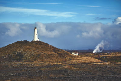 White Reykjanesviti lighthouse standing on a low hill with geothermal steam in the distance on the Reykjanes Peninsula, Iceland.
