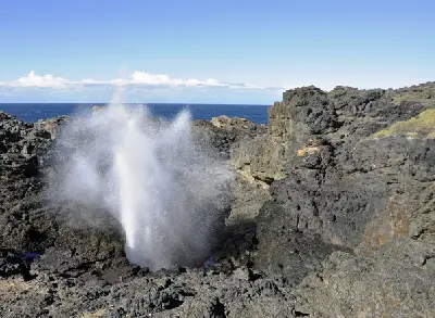 Powerful jet of seawater erupts from the Kiama Blowhole amid rugged dark coastal rocks, with the blue Pacific Ocean beyond.