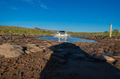 Four-wheel-drive vehicle splashes through the rocky shallows of the Pentecost River with the Cockburn Range in the distance.