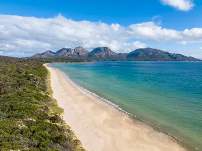 Aerial view along Muirs Beach toward the rugged granite peaks of The Hazards across blue-green waters near Coles Bay, Tasmania.