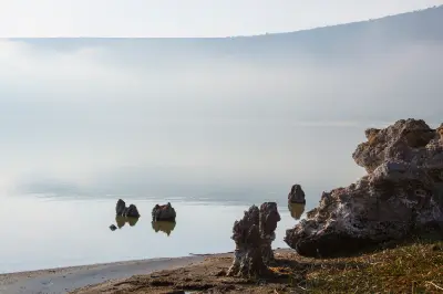 Morning light and mist over Empakaai Crater Lake in northern Tanzania, with calm reflective water, volcanic rocks along the shoreline and the forested crater rim in the background.