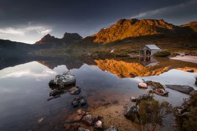 Dove Lake and its historic boatshed reflecting in calm water beneath sunlit mountains in Tasmania, Australia.