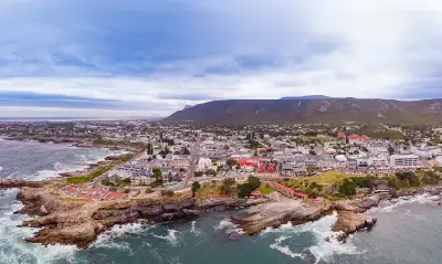 Coastal aerial panorama of Hermanus with rocky ocean cliffs, shoreline buildings, and mountains along the Whale Coast in South Africa.