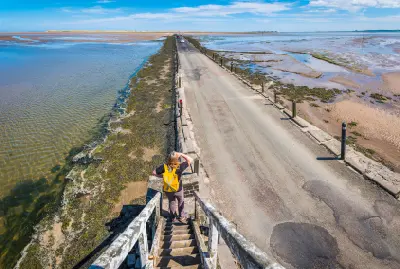 Elevated view of the Holy Island Causeway crossing extensive tidal sands and shallow water in Northumberland, with a woman standing on steps overlooking the road.