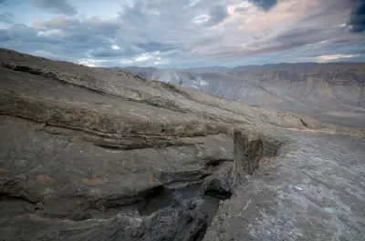 Rocky volcanic landscape on the upper slopes of Ol Doinyo Lengai, with lava formations, eroded terrain and the Great Rift Valley stretching into the distance under a dramatic sunrise sky.
