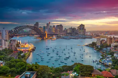 Elevated sunset view across Sydney Harbour with the Harbour Bridge, illuminated CBD skyline, Luna Park and yachts anchored in Lavender Bay.