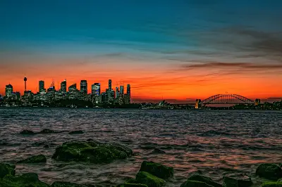 Fiery orange sunset over Sydney Harbour from Bradleys Head, with the CBD skyline, Opera House and Harbour Bridge beyond the rocky shoreline.