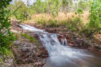 Flowing white water cascading over reddish rock ledges at Buley Rockhole, surrounded by tropical woodland in Litchfield National Park, Northern Territory, Australia.