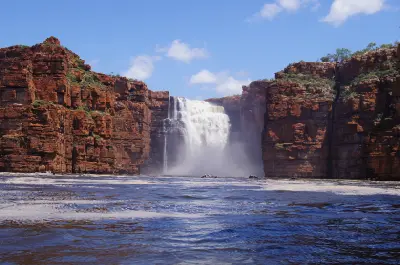 Powerful King George Falls plunges from towering red sandstone cliffs into the tidal gorge, with small boats below showing the immense scale.