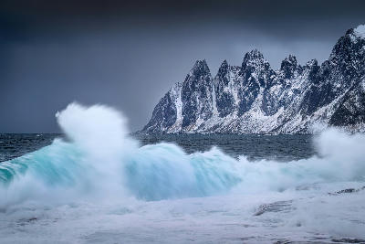 Waves crashing in foreground with Okshornan Devil’s Teeth peaks, Senja Norway, snow covered jagged mountains along the coast at Tungeneset.