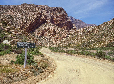 Entrance sign and gravel road leading into Seweweekspoort beneath folded sandstone mountains in the Klein Karoo, South Africa.