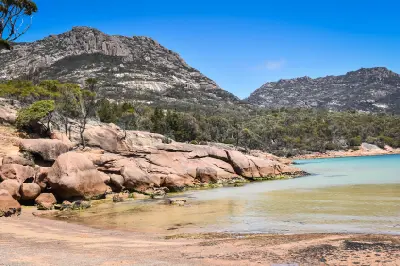 Pink granite rocks and shallow turquoise water at Honeymoon Bay with the rugged peaks of The Hazards rising behind, Freycinet National Park, Tasmania.