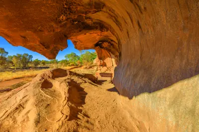Wave-shaped sandstone overhang along the Mala Walk at the base of Uluru, with red rock formations and Outback vegetation beyond.