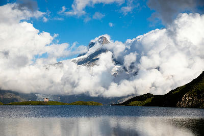 Cloud-wrapped alpine peak rising above Bachalpsee with calm water reflections and a small hut on the lakeshore, Bernese Alps, Switzerland.