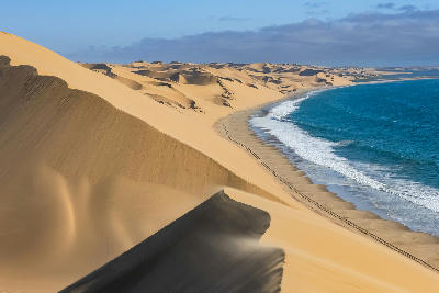 Large sand dunes meeting the Atlantic shoreline along Namibia’s Skeleton Coast with ocean surf and wind-blown desert sand.