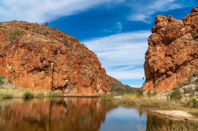 Towering, massive red-orange cliffs frame a peaceful waterhole on the Finke River with thick green reeds growing along the water's edge.
