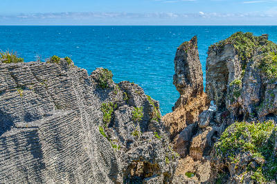 Layered grey limestone stacks of the Pancake Rocks at Punakaiki, with a tall sea stack and green coastal plants above the deep blue Tasman Sea, Paparoa National Park, New Zealand.