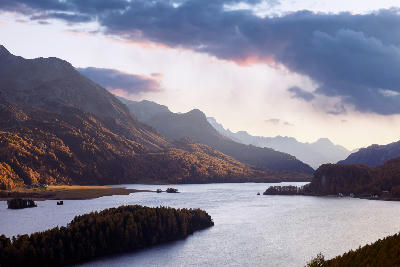 Lake Sils in autumn with golden larch forests, small islands, and surrounding mountain ridges in the Upper Engadin region of Switzerland.