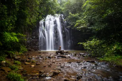 Ellinjaa Falls surrounded by dense tropical rainforest, with white cascades descending into a pool and a rocky stream in the foreground.