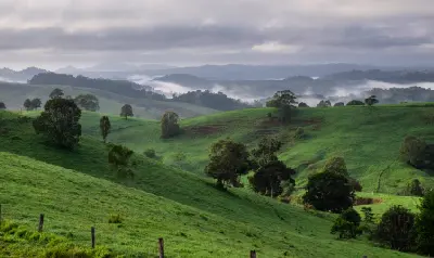 Rolling green hills and scattered trees overlooking misty valleys and distant mountains in the Atherton Tablelands, Queensland.