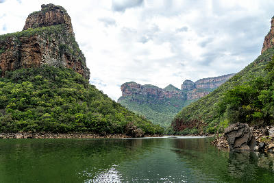 View from a boat on the Blyde River within Blyde River Canyon, showing steep canyon walls, lush vegetation, calm water and the Three Rondavels rising above the landscape in Mpumalanga, South Africa.