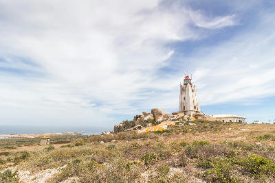 Cape Columbine Lighthouse standing on a rocky headland above coastal vegetation with the Atlantic Ocean visible in the background near Paternoster, South Africa.