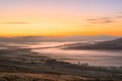Aerial sunrise view of Ribblehead Viaduct stretching across mist-filled valleys beneath an orange and pink sky in the Yorkshire Dales, England.