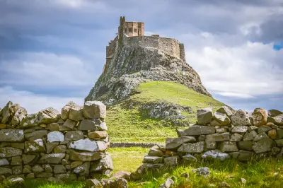 Lindisfarne Castle perched on a steep rocky outcrop above a green hillside, framed by old dry-stone walls beneath a cloudy sky on Holy Island, Northumberland.