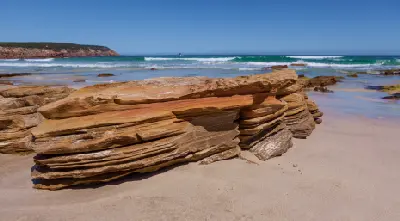 Close-up of layered orange sandstone rocks on the sandy shoreline of Stokes Bay, Kangaroo Island, with ocean waves in the background.