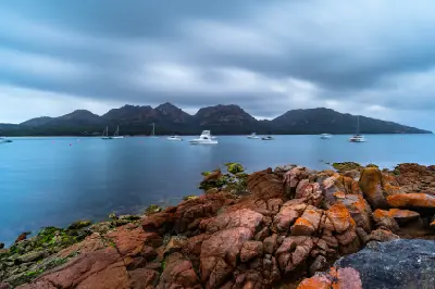 The Hazards mountain range viewed across Coles Bay, with boats on calm blue water and orange-lichen-covered granite rocks in the foreground, Tasmania.